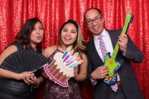 Two women and a man smiling in a photobooth with a red sequin backdrop, holding fun props like a birthday cake cutout, a fan, and a toy guitar.