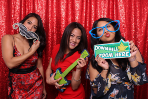 Three women posing in a photobooth with playful props, including a fake mustache, a toy guitar, and oversized glasses. The backdrop is a red sequin curtain.