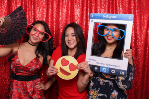 Three women in fun oversized props posing in a photobooth with a red sequin backdrop. One holds a fan, another an emoji face, and the third a large Instagram frame.