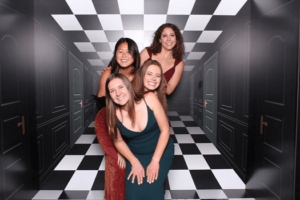 Four friends posing playfully in elegant dresses against a checkered 3D illusion backdrop featuring a surreal hallway with multiple doors.